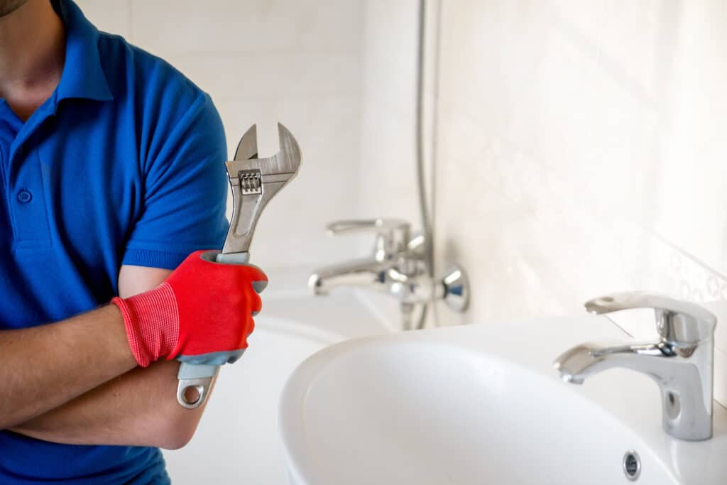 Plombier en polo bleu et gant rouge tenant une clé à molette, prêt à réparer un évier de salle de bain avec robinet chromé.