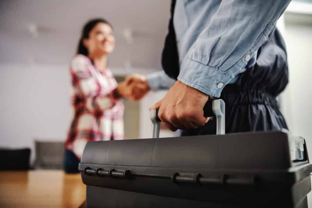 Main d'un réparateur en chemise denim tenant une boîte à outils, serrant la main d'une cliente souriante en arrière-plan flou, signe de satisfaction.