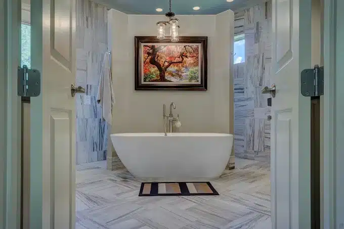 Salle de bain luxueuse et moderne avec baignoire îlot blanche, carrelage marbre gris, grand tableau d'arbre coloré et portes ouvertes.